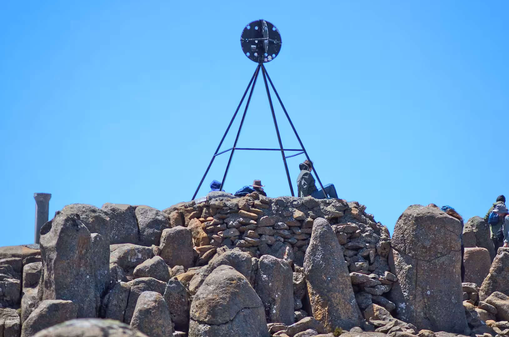Hikers rest on the stone cairn beneath a steel tripod marker at a rocky mountain summit.