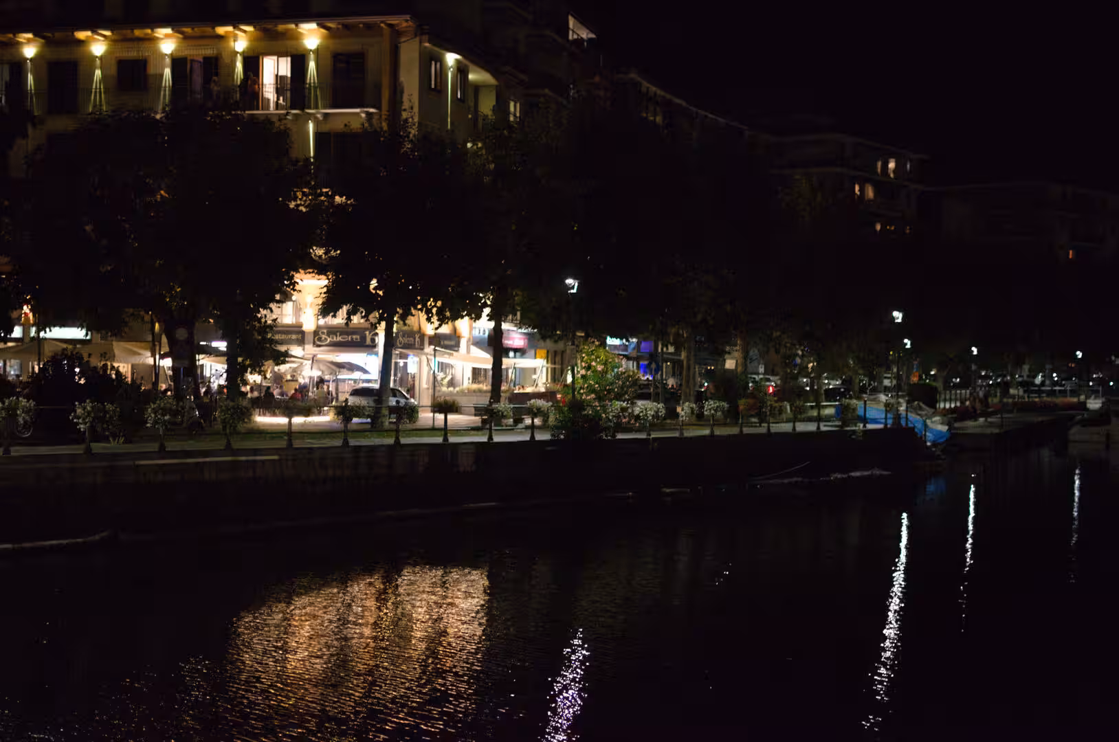A warm glow of cafe awnings and street lamps reflects across the dark water on a quiet waterfront.