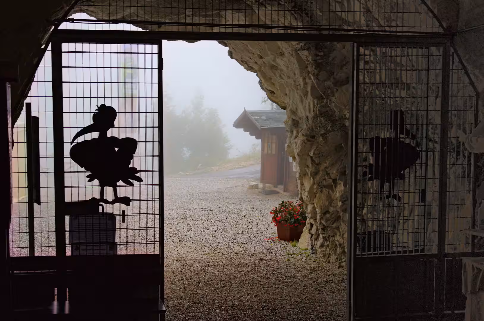 Looking out from a grotto's wire gate, stork figures stand in silhouette against swirling fog and a small wooden hut.