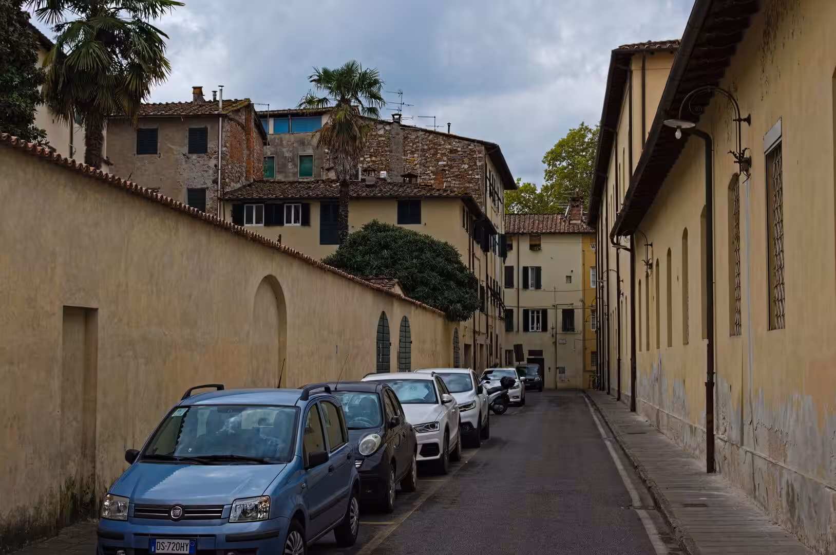 A row of parked cars lines a curving lane between aged stucco and brick walls.