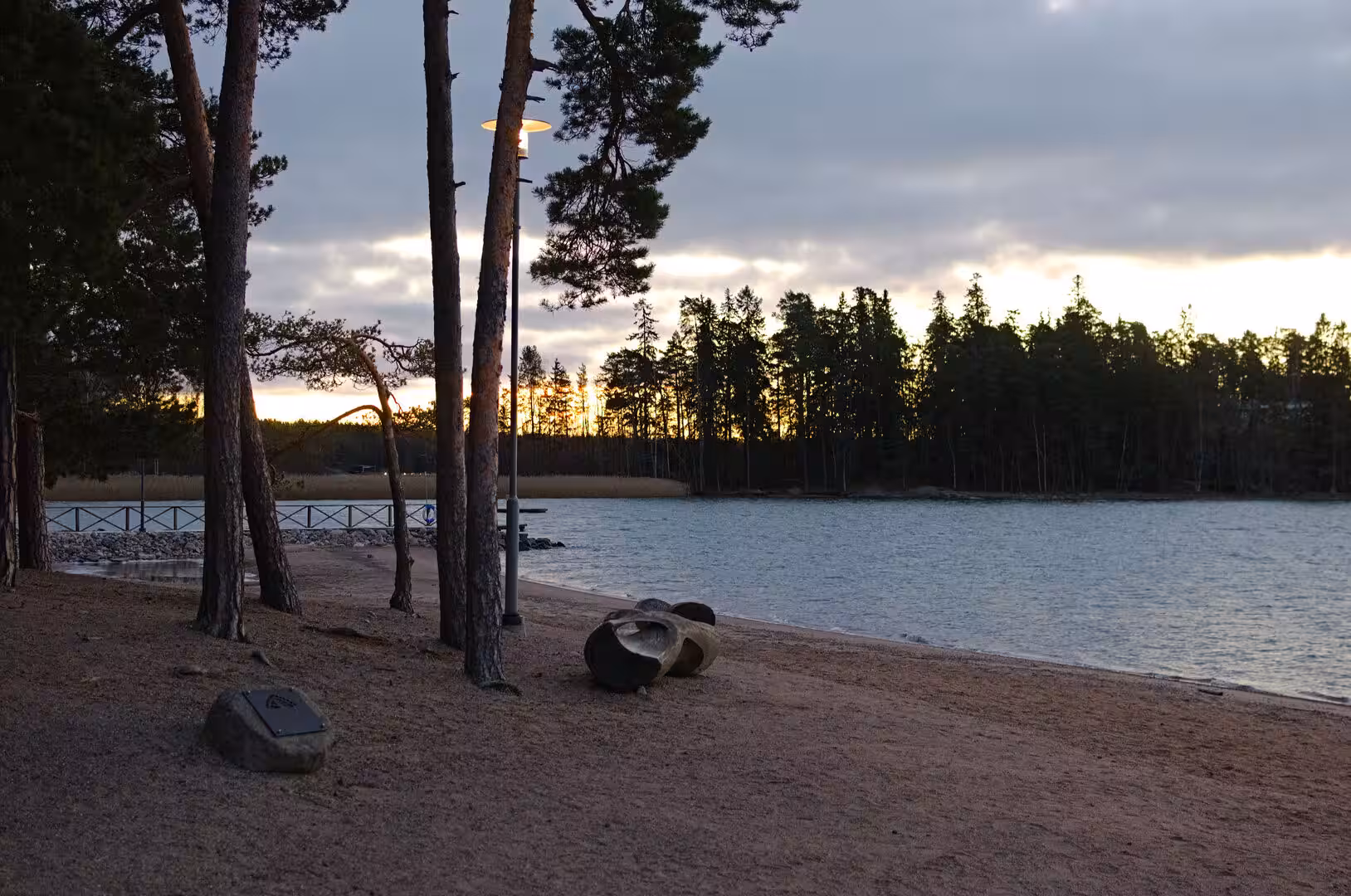 A boulder and a low marker sit on the sand as the sun sets behind the treeline.