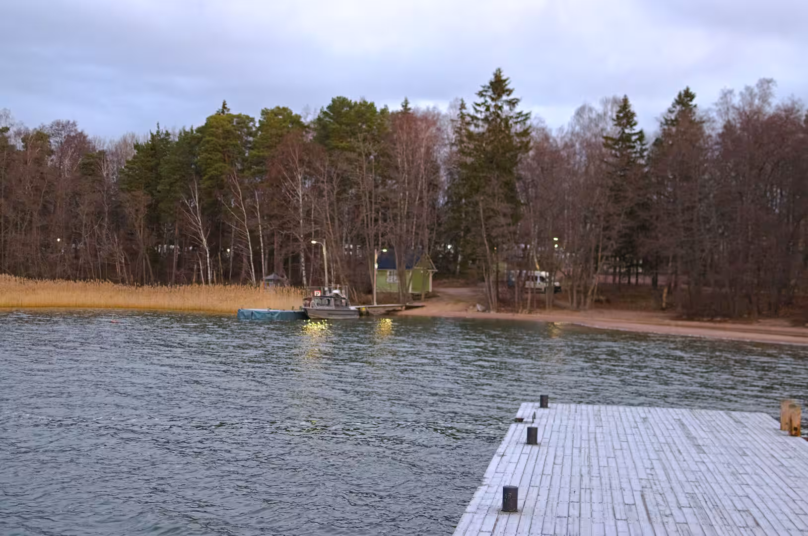 A weathered wooden pier reaches out toward a boat moored by a green cabin among bare trees.
