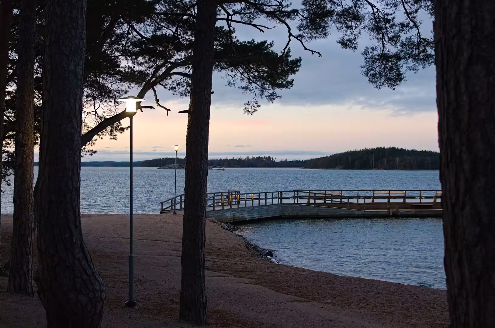 A lamplit wooden jetty extends from a sandy beach into calm water, seen through pine trunks.
