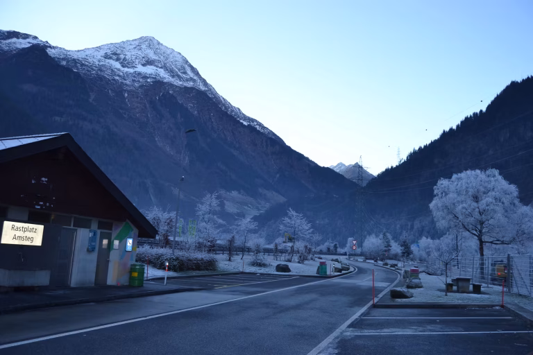 A quiet roadside rest area beside a building with a German rest-stop sign, snow-capped peaks towering behind, and frost-covered trees lining the empty parking lot.