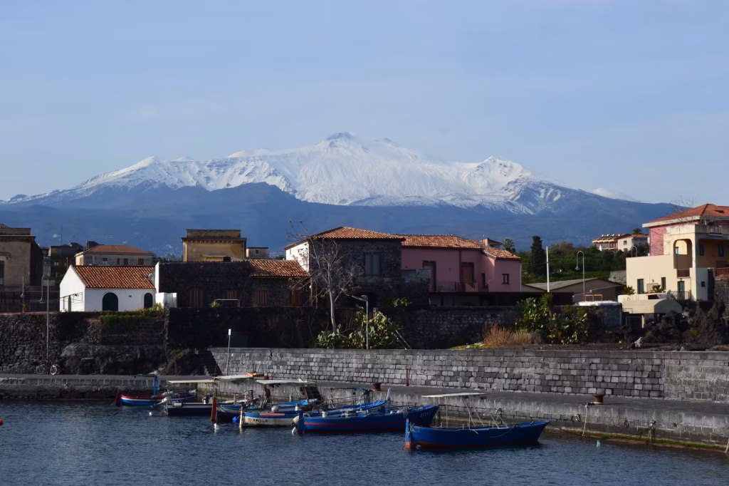 Small fishing boats moored in a harbor beside a stone quay and village houses, with snow-capped Mount Etna rising behind.