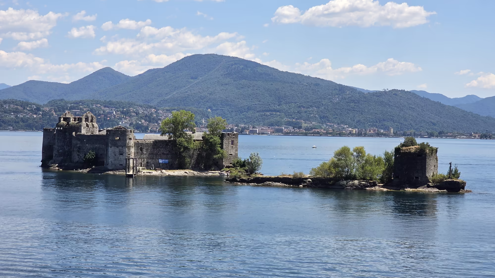 A ruined stone fortress and a small round tower stand on an island in the lake near Cannobio, with forested mountains rising behind.