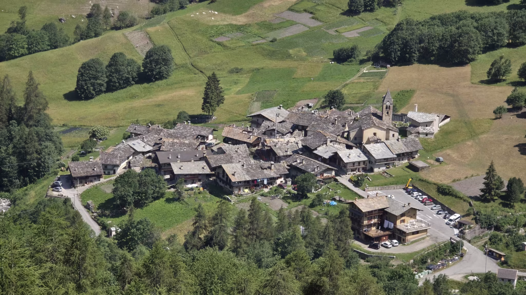 A cluster of alpine houses with grey slab roofs surrounds a church bell tower on a green slope above the valley.