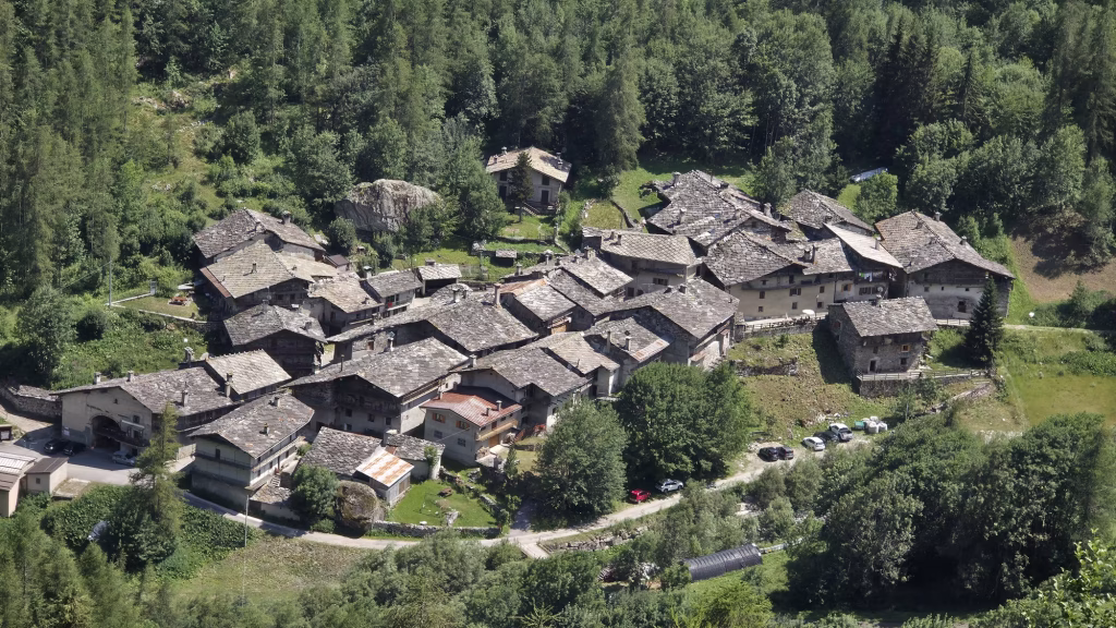 A tight group of stone houses with slab and metal roofs sits on a terrace surrounded by dense woodland.