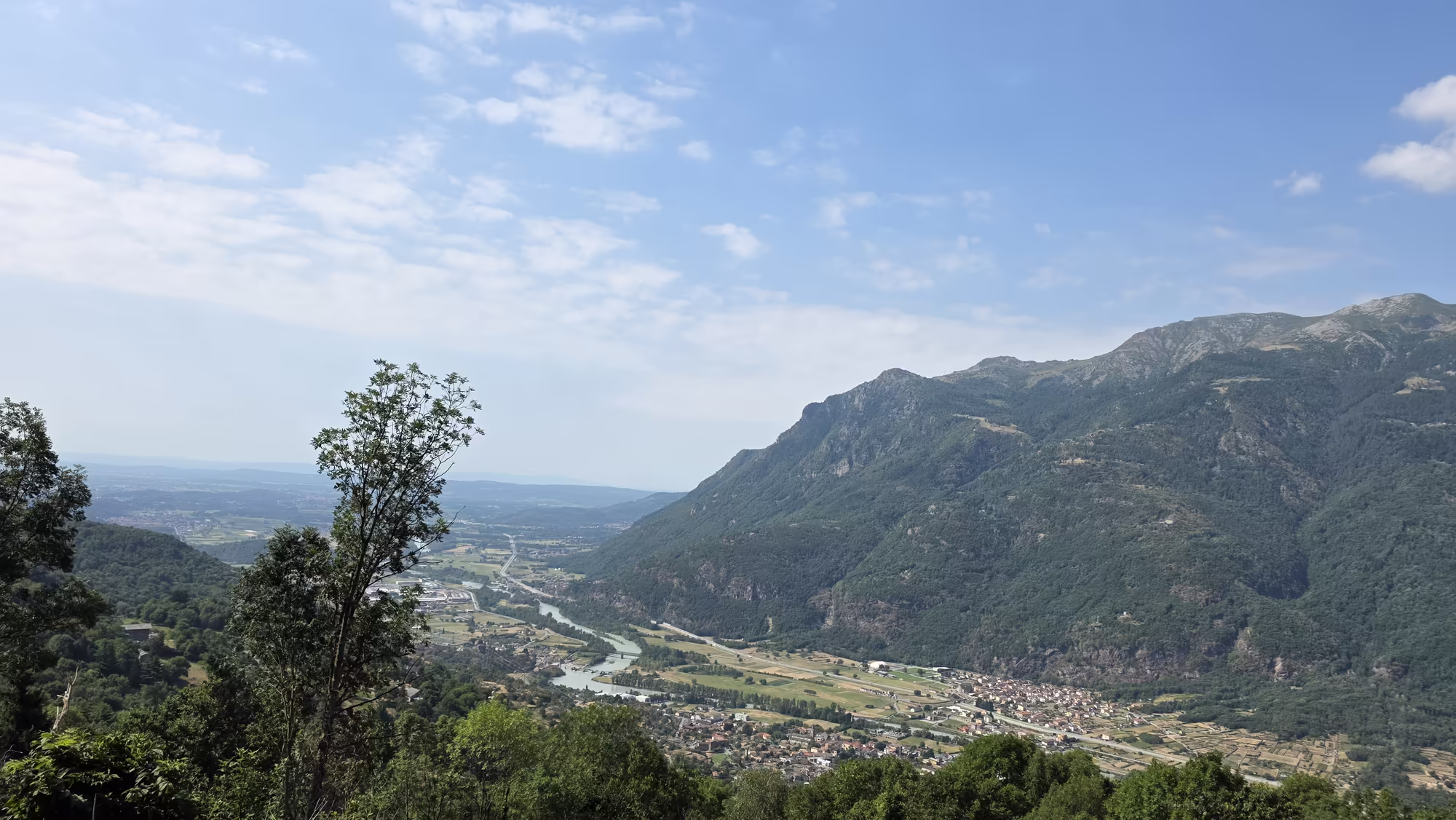 A broad river valley with a meandering river, fields and a small town is seen from a wooded height near Settimo Vittone in Piedmont.