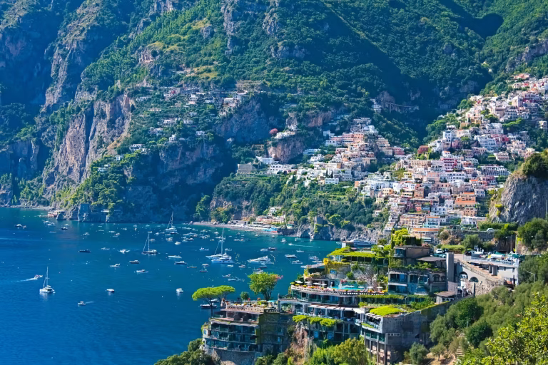 A dense village of white buildings clings to steep, green mountainsides overlooking a deep blue bay filled with boats.