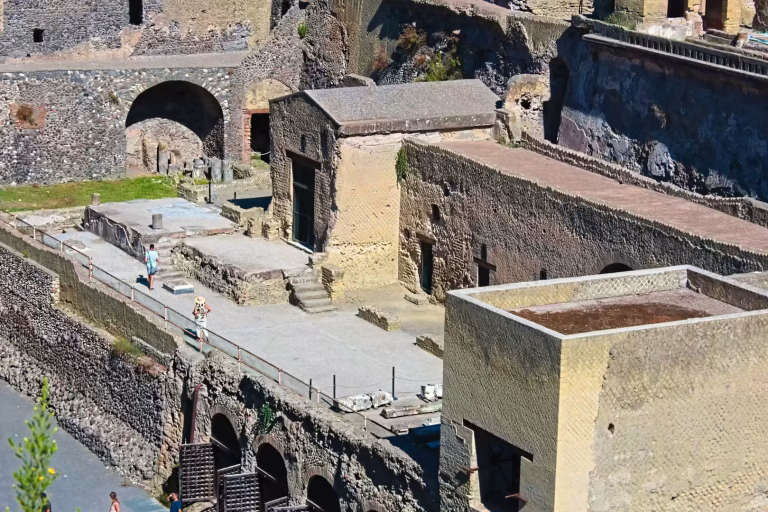 A view of ancient stone ruins with arched openings, a small building with a pitched roof, and a paved walkway with a few people.
