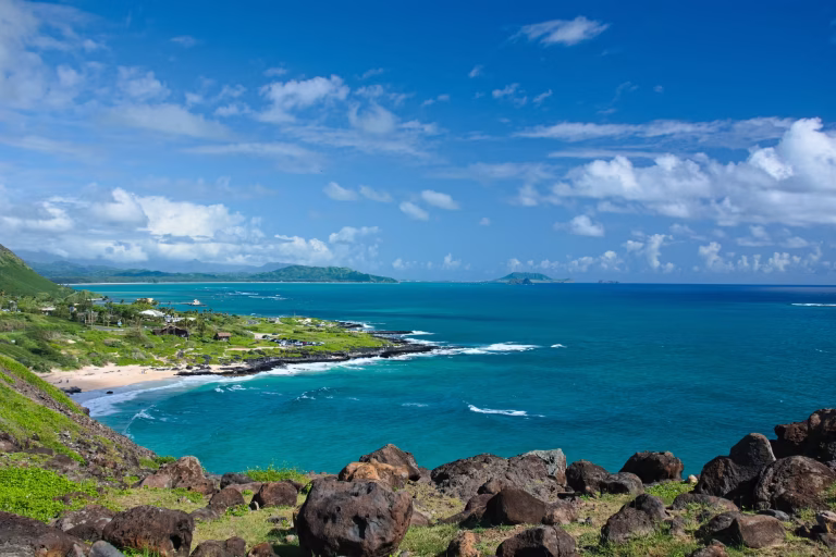 Heavy dark boulders lie scattered across a grassy bank above the Makapuʻu shoreline, with white surf breaking on the reef below.