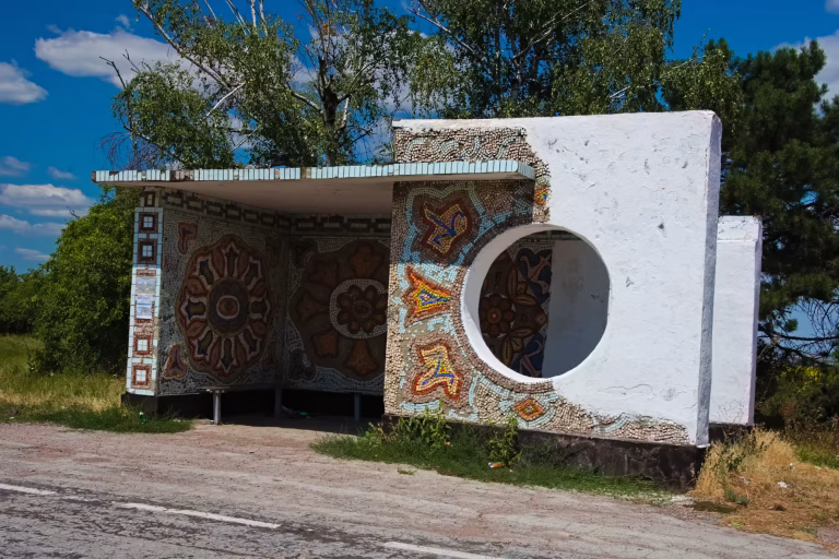 A white concrete bus shelter features a large circular opening and intricate geometric mosaics in earthy brown and blue tones.