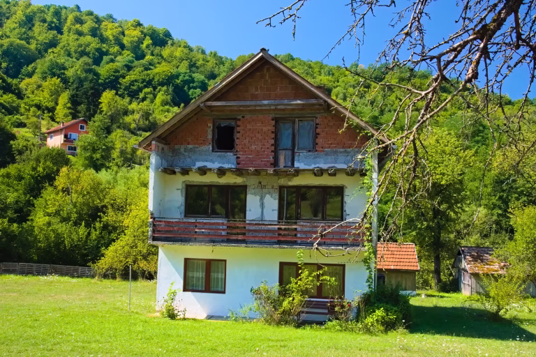 A part-finished two-storey house with a white plastered lower floor, exposed brick upper storey and wooden balcony railings stands on a grassy slope below a densely wooded hill.