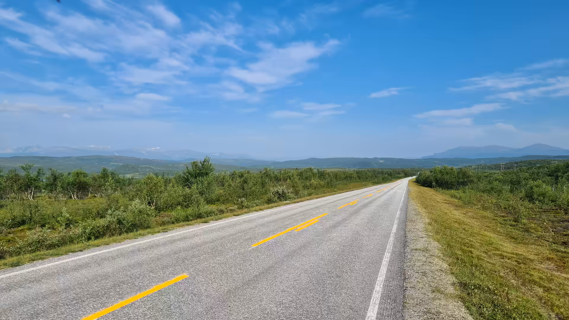 A grey asphalt road curves through open green countryside beneath a bright blue sky streaked with thin white clouds, at Lakselv in Finnmark, Norway.