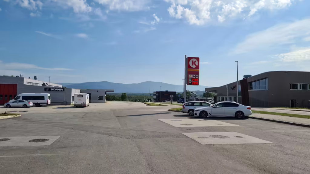 A tall fuel price sign stands on grey asphalt before industrial buildings, one of them bearing the word SCANIA, with mountains behind, at Lakselv in Finnmark, Norway.
