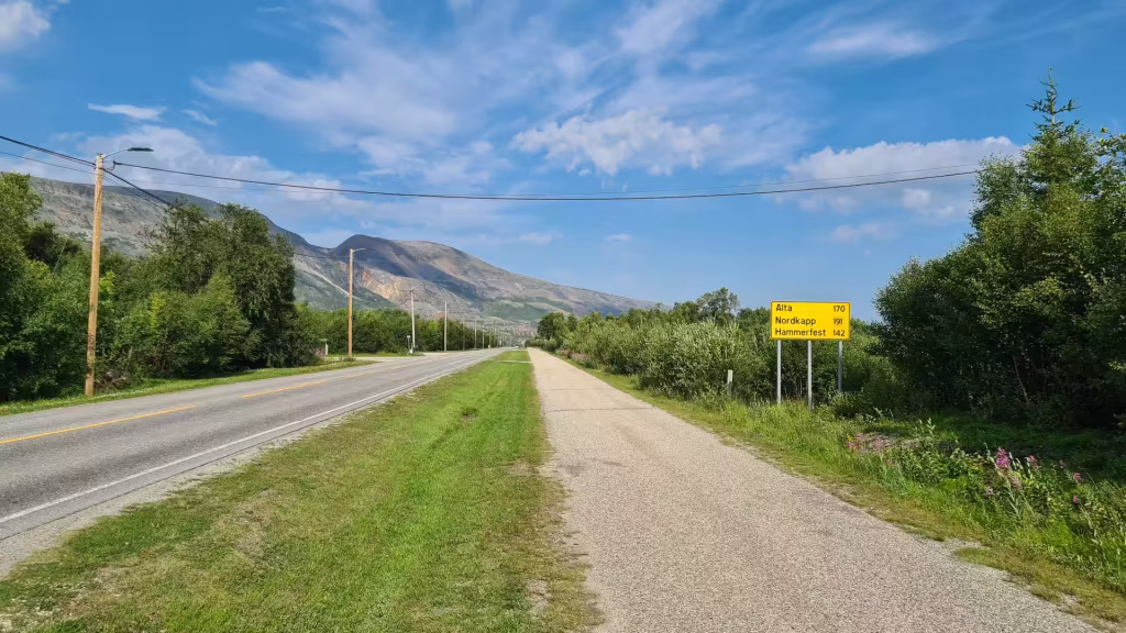A yellow sign listing the distances to Alta, Nordkapp and Hammerfest stands beside a paved road and gravel path below rocky hills, at Lakselv in Finnmark, Norway.