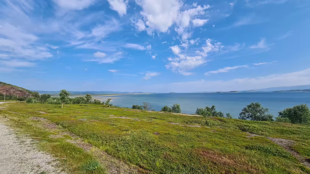 A wide grassy field slopes gently toward a large blue expanse of water under a bright sky of puffy white clouds, at Lakselv in Finnmark, Norway.