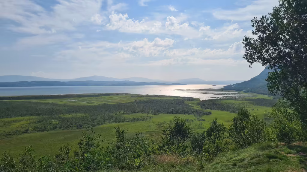 An elevated view looks across a flat green plain and a large pale stretch of water toward low hazy hills on the horizon, in Porsanger in Finnmark, Norway.