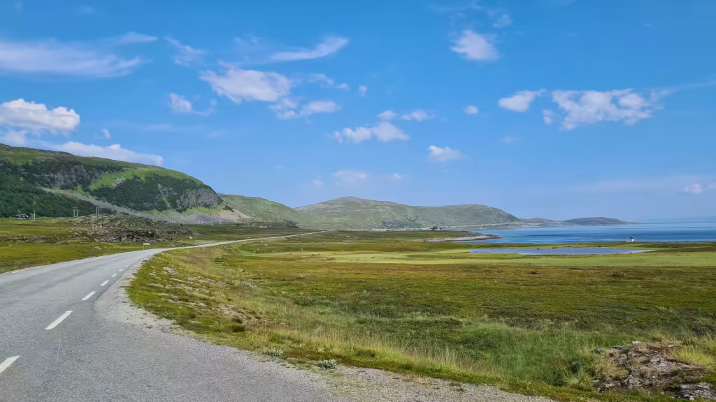 A paved road marked with a white dashed line curves through green countryside beside a wide marshy plain, water and distant hills, at Kafjord in Finnmark, Norway.