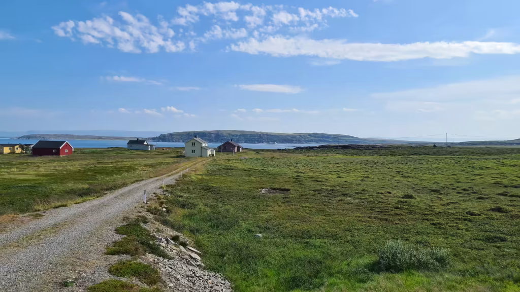 A light gravel track runs through open grassy ground past a few small houses toward blue water and a long low ridge, at Kafjord in Finnmark, Norway.