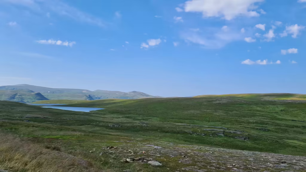 Green and yellow grass rolls across an open landscape with a narrow blue lake lying in a hollow below rounded hills, at Kafjord in Finnmark, Norway.