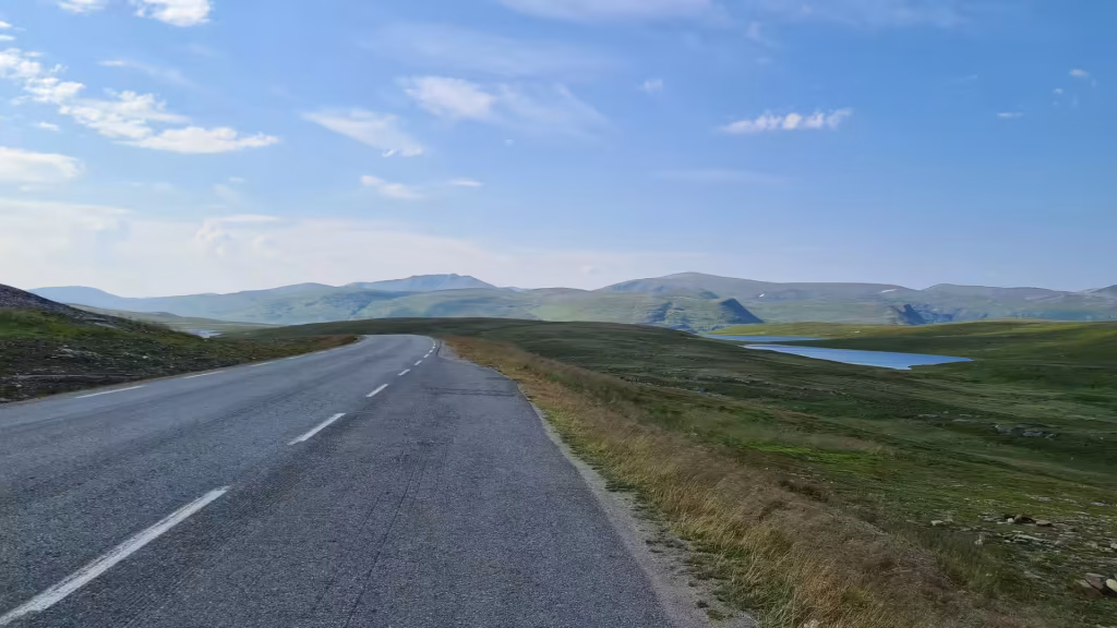 A grey road with a white dashed line curves through treeless upland terrain beside a blue lake and distant rolling hills, at Kafjord in Finnmark, Norway.