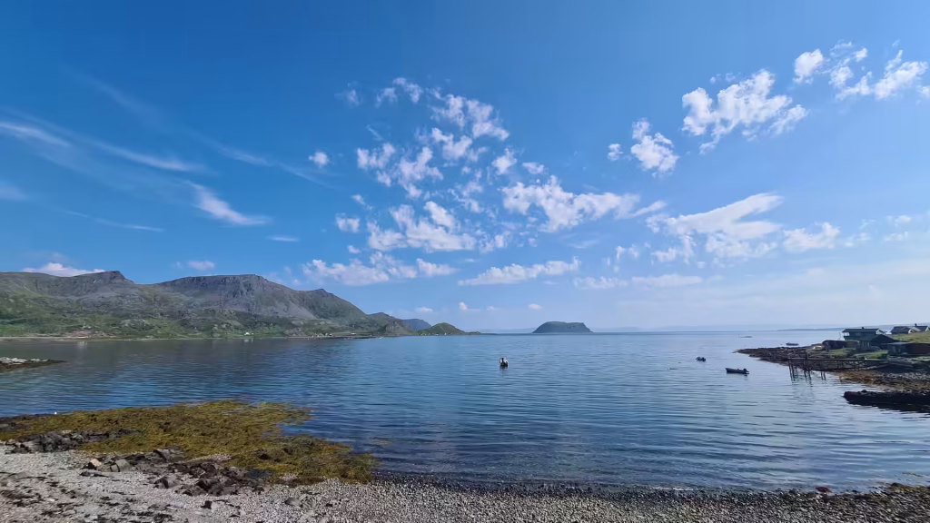 A pebbled shore meets calm water below a rugged mountain ridge, with small boats and a low shoreline of buildings beyond, at the North Cape in Finnmark, Norway.