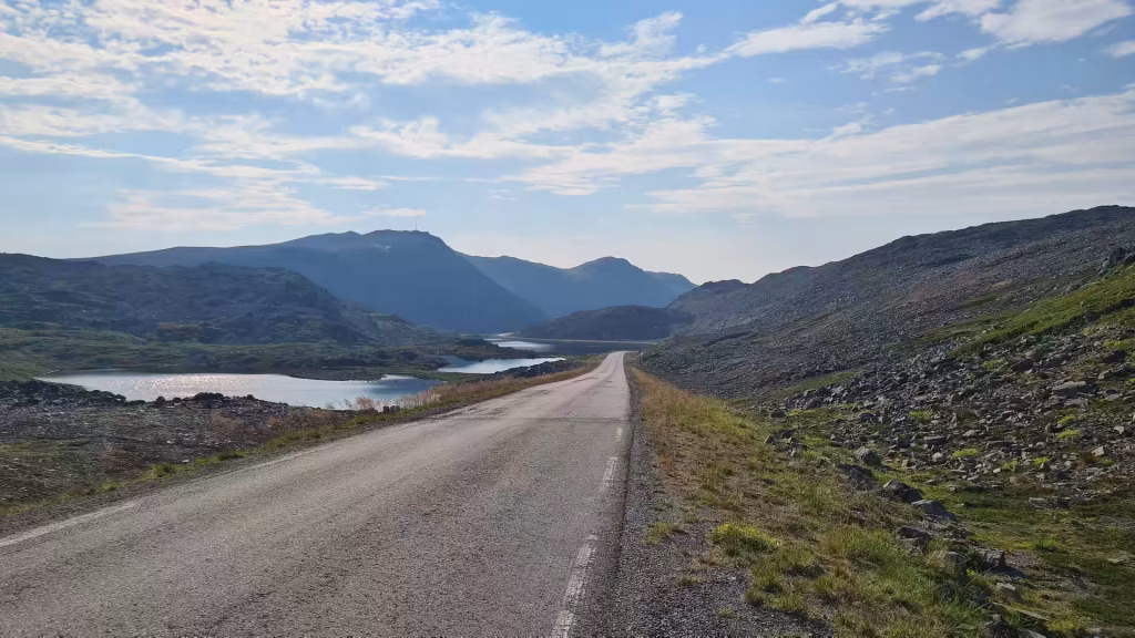 A grey road with a white dashed line runs through bare rocky hills past small lakes and rounded mountain ridges under a clouded sky, at Gjesvaer on Mageroya in Finnmark, Norway.