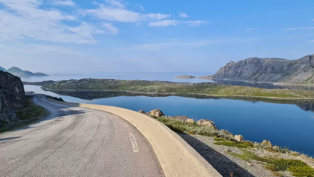 A grey road sweeps around a low concrete barrier on a rocky slope above calm water dotted with small islands, at Gjesvaer on Mageroya in Finnmark, Norway.