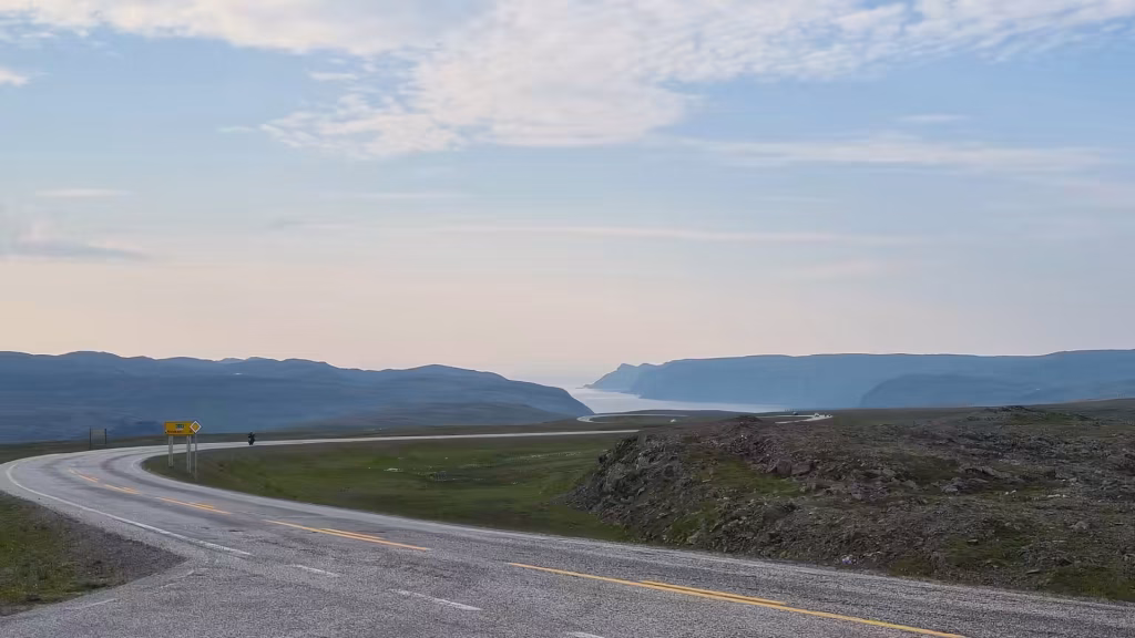 An asphalt road marked with yellow centre lines sweeps past a small warning sign towards blue hills and water, at the North Cape in Finnmark, Norway.
