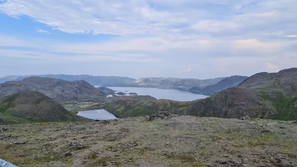 Rugged rock and moss cover the foreground ledge above a wide lake ringed by layered hazy mountains, at the North Cape in Finnmark, Norway.