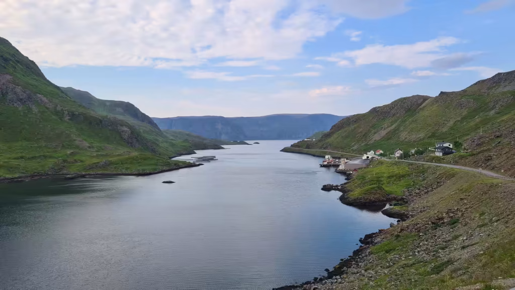 A calm inlet lies between steep green slopes with a few small buildings dotted along the right shore, at Kamoyvaer on Mageroya in Finnmark, Norway.