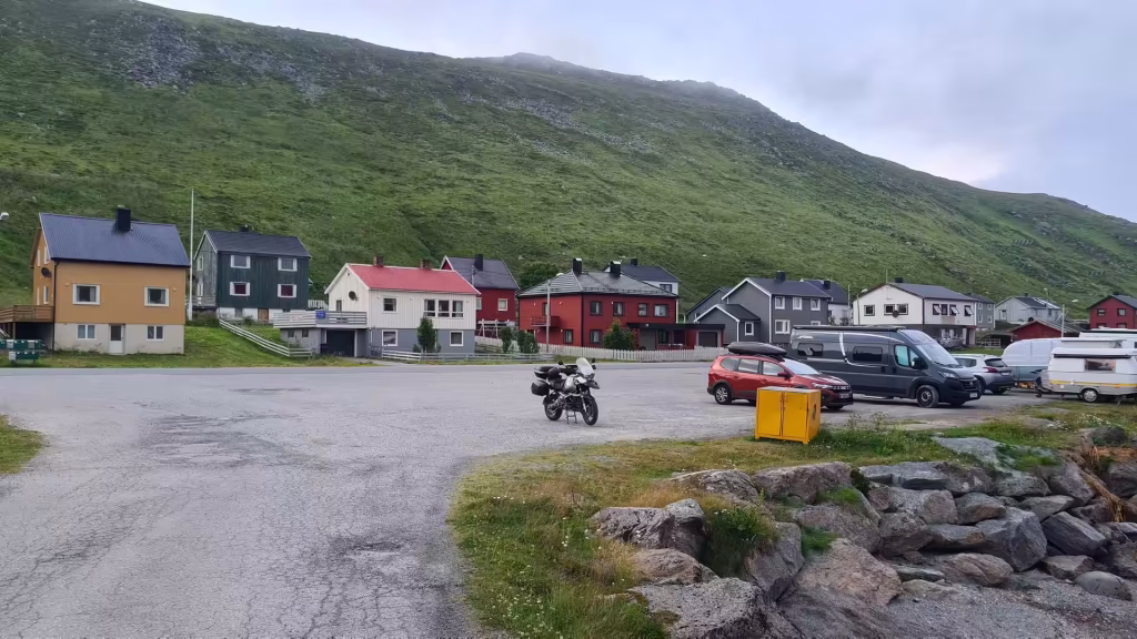 A loaded BMW GS adventure motorcycle stands on cracked asphalt facing a row of houses below a steep green slope, at Skarsvag on Mageroya in Finnmark, Norway.