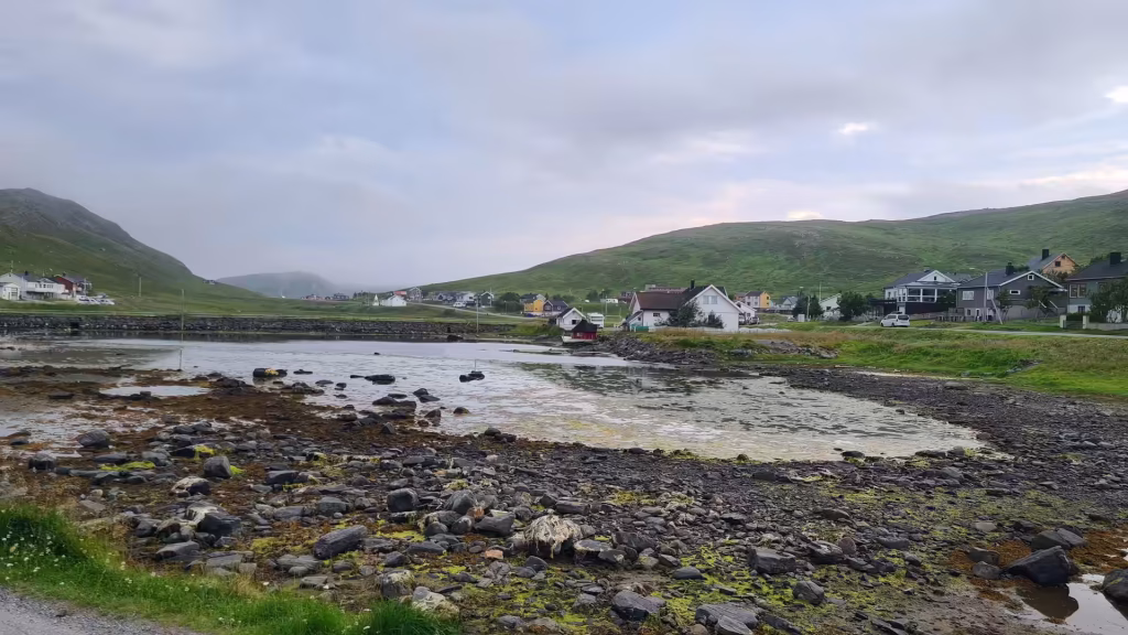 Dark wet rocks and seaweed lie exposed in the foreground of a shallow bay below a scattered coastal village, at Skarsvag on Mageroya in Finnmark, Norway.