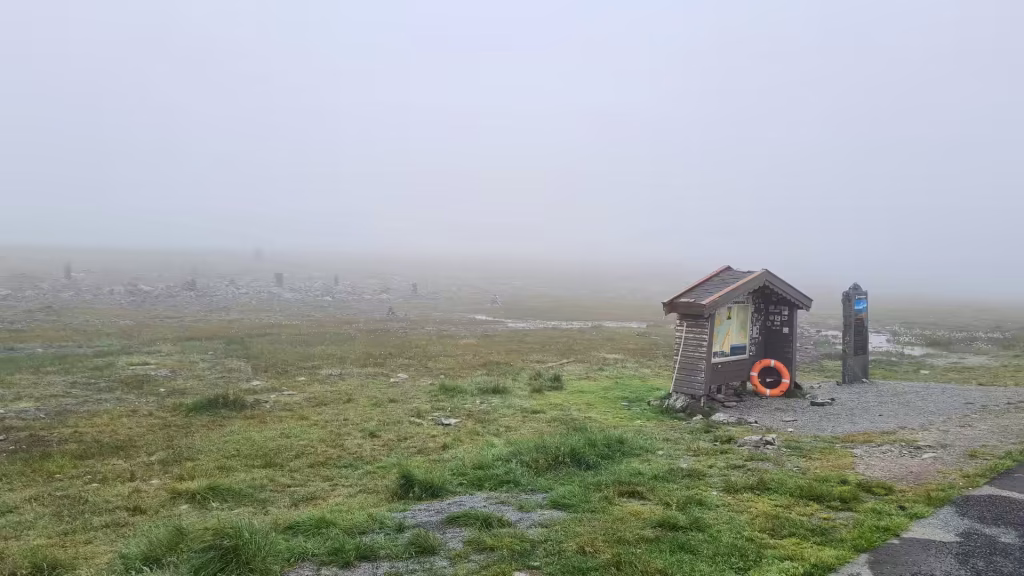 A small weathered wooden hut with an orange lifebuoy stands on wet gravel beside a stone marker in thick grey fog, at the North Cape in Finnmark, Norway.