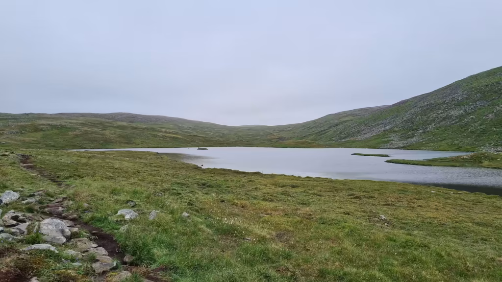 A calm grey lake lies between rounded grassy hills with a rocky path crossing the foreground grass, at the North Cape in Finnmark, Norway.