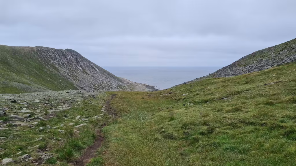 A worn dirt path climbs a grassy col between two slopes and opens onto a wide grey sea under an overcast sky, at the North Cape in Finnmark, Norway.