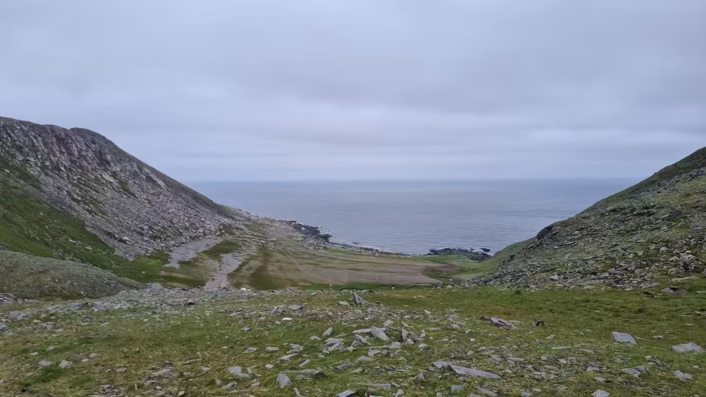 A grassy valley with a faint track on its left slope falls to a rocky shore beside a calm grey sea, at the North Cape in Finnmark, Norway.