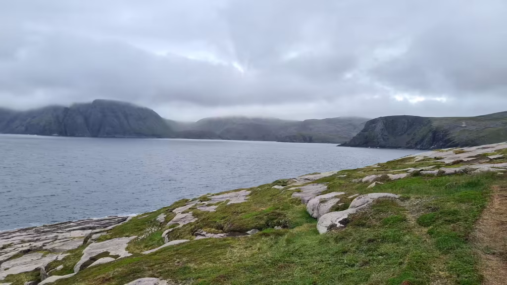 Flat grey rock slabs and green grass edge a calm sea that stretches toward distant headlands under layered cloud, at the North Cape in Finnmark, Norway.