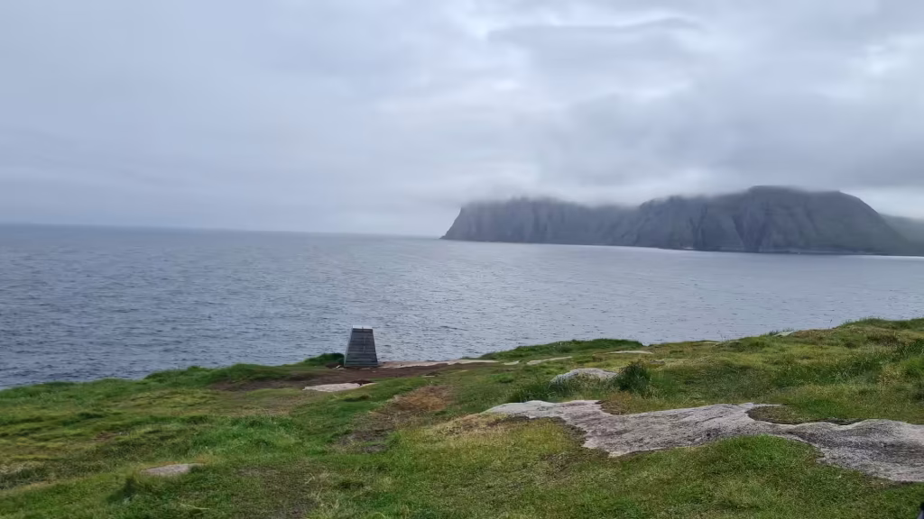 A low dark marker stands on a grassy cliff edge overlooking grey water and a fog shrouded rock mass, at the North Cape in Finnmark, Norway.