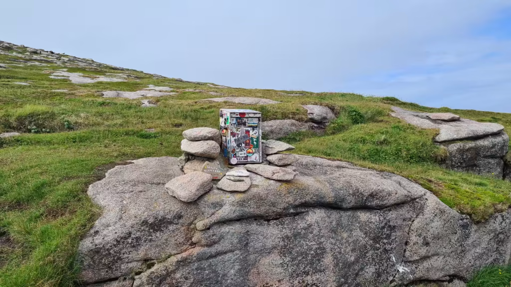 A small white box covered in travel stickers sits on bare rock beside a wooden honesty box sign in open country, at the North Cape in Finnmark, Norway.