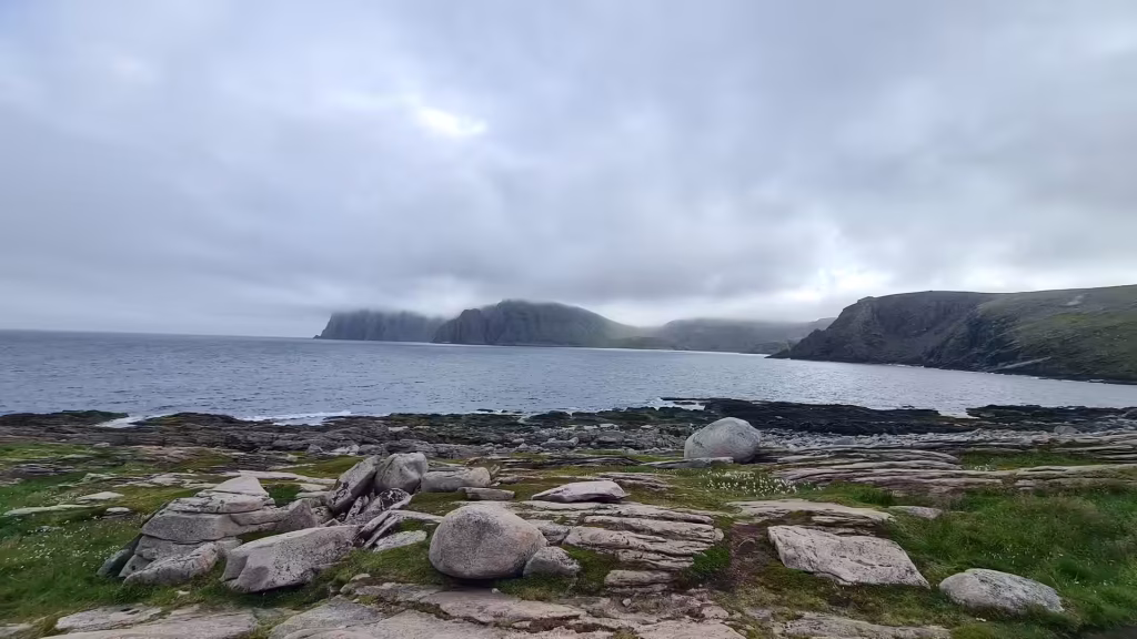 Layered grey bedrock and boulders lead to calm water beneath a dark cliff hidden in low cloud, at the North Cape in Finnmark, Norway.