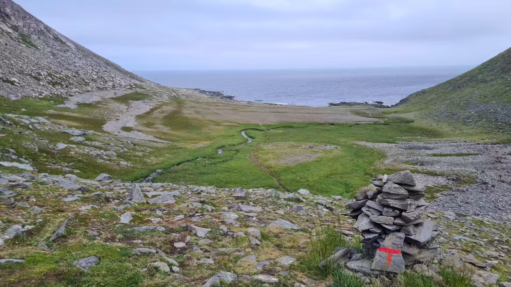 A stacked stone cairn marked with red paint stands on rocky ground above a green valley opening to the sea, at the North Cape in Finnmark, Norway.