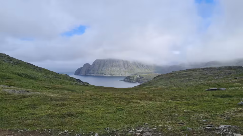 Short grass and scattered stones slope down to dark water and a steep cliff wrapped in low cloud, at the North Cape in Finnmark, Norway.