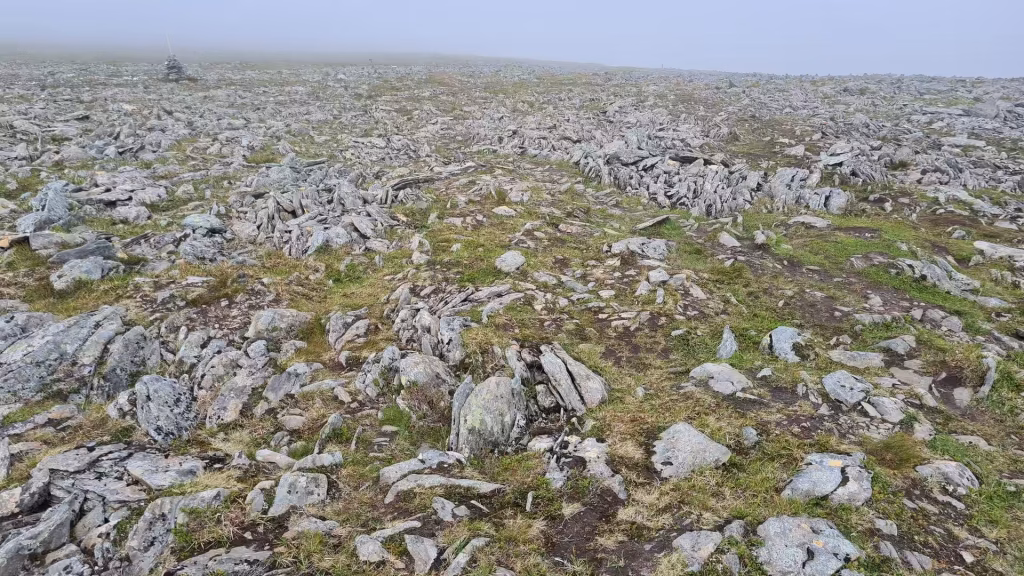Grey fractured stones and low mossy plants cover the ground while a faint cairn and post vanish into mist on the horizon, at the North Cape in Finnmark, Norway.