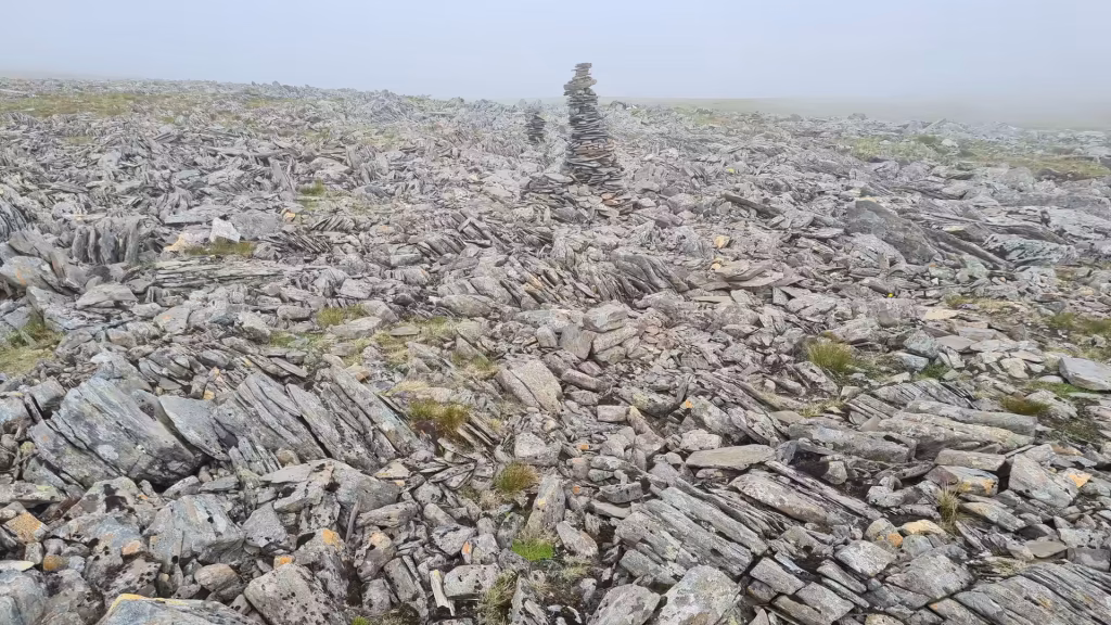 A hand built stack of flat stones rises from barren rocky ground that fades into grey mist with no horizon visible, at the North Cape in Finnmark, Norway.