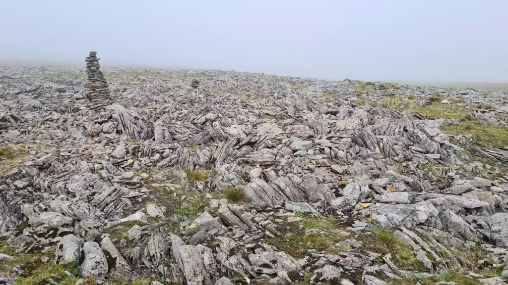 A cairn of stacked flat stones stands on a rocky, grassy plateau shrouded in thick fog, at the North Cape in Finnmark, Norway.