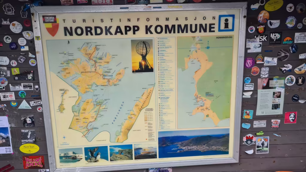 A framed tourist information board with maps and photographs hangs on a dark wall covered in stickers, at the North Cape in Finnmark, Norway.