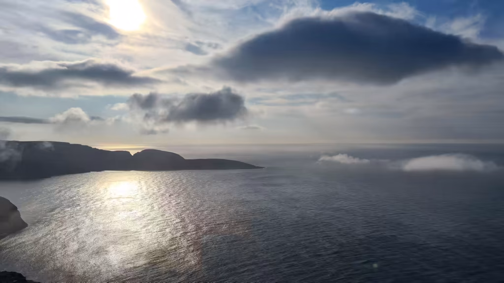 Sunlight glares through thin cloud above a rippled sea with dark headlands and a low fog bank, at the North Cape in Finnmark, Norway.